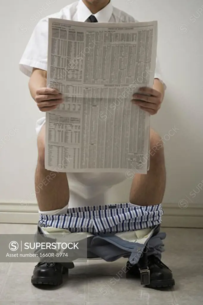 Mid section view of a young man sitting on the toilet seat reading a newspaper