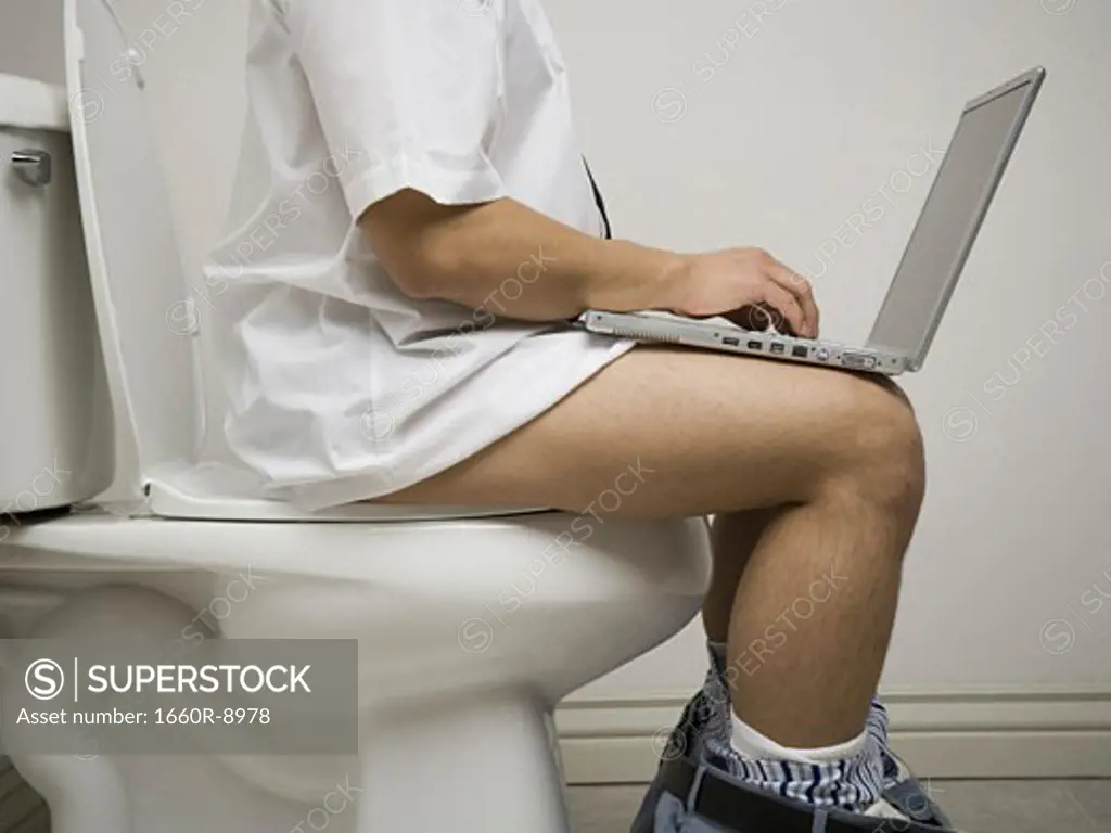 Mid section view of a young man sitting on the toilet seat using a laptop