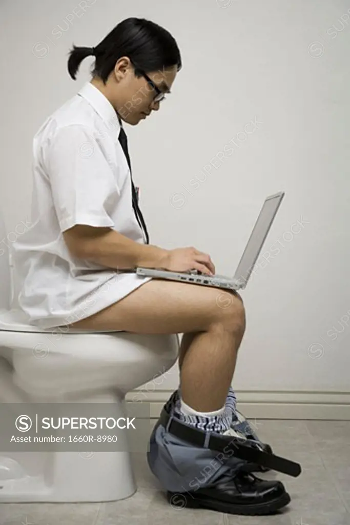 Profile of a young man sitting on a toilet using a laptop