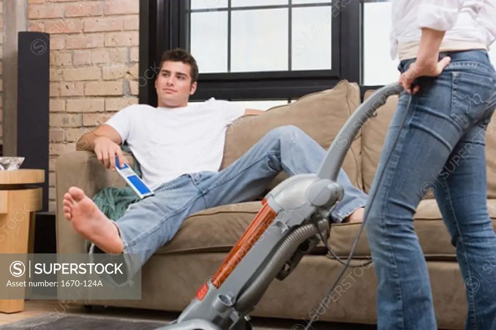 Teenage boy sitting on a couch with a woman vacuuming the floor