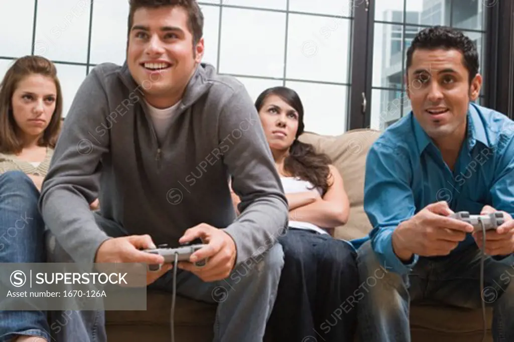 Young man and a teenage boy playing a video game with two young women sitting beside them looking displeased