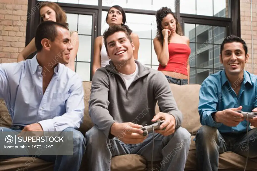 Young man and a teenage boy playing a video game with three young women standing behind them looking displeased