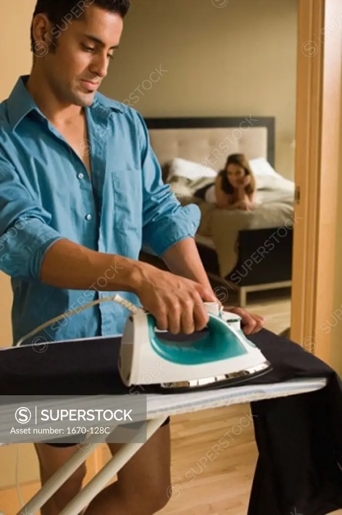 Close-up of a young man ironing clothes with a young woman lying on the bed in the background