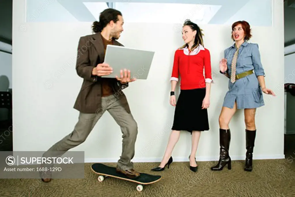 Businessman holding a laptop and riding a skateboard past two businesswomen
