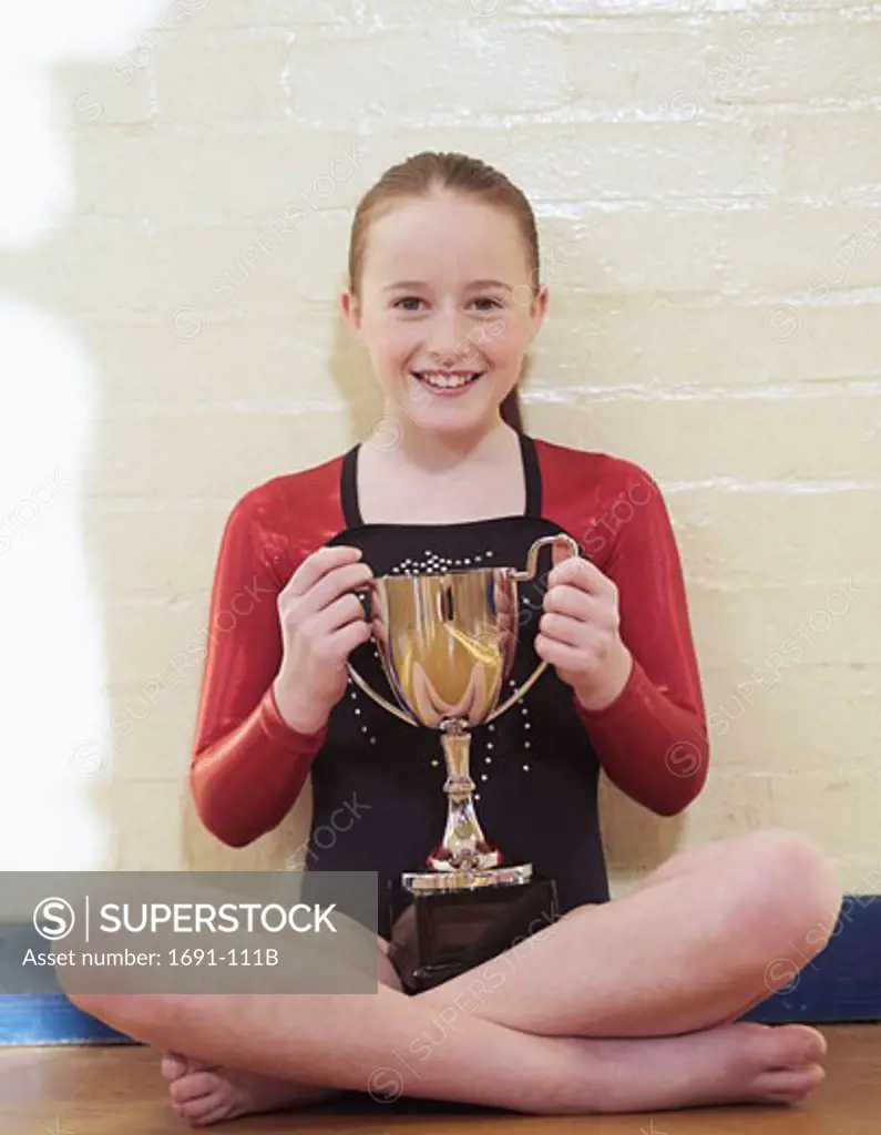 Female gymnast sitting by a wall and holding a trophy