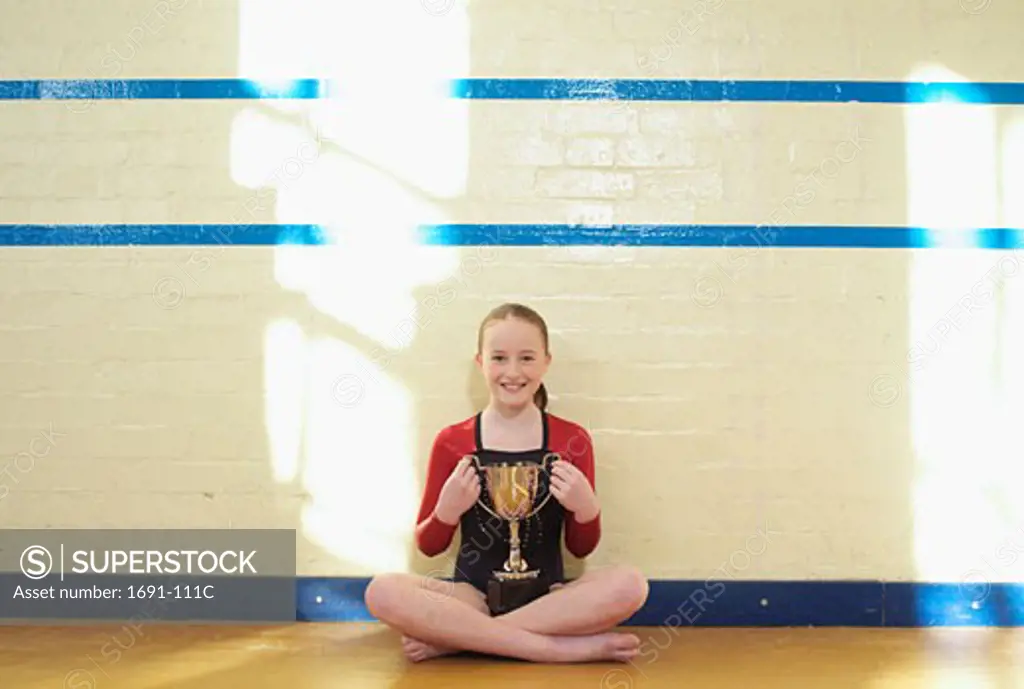 Female gymnast sitting by a wall and holding a trophy