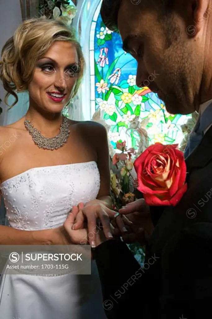 Groom putting a ring on a bride's finger in front of a stained glass window