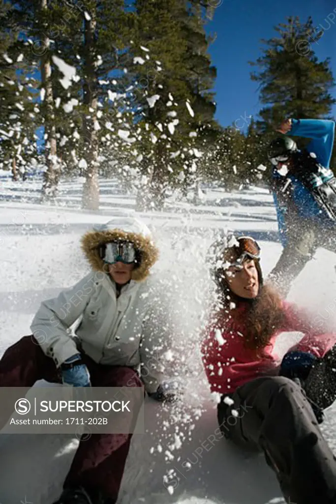 Two young woman sitting on snow with a mid adult man snowboarding behind them