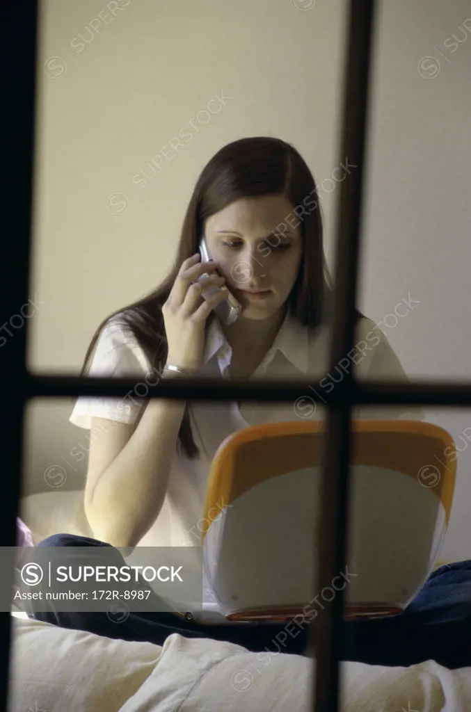 Teenage girl sitting on a bed in front of a laptop talking on a mobile phone