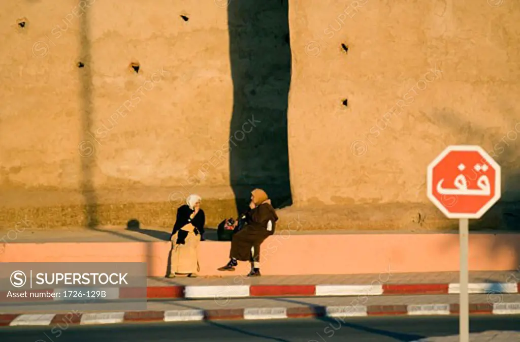 Two people sitting on the ledge of a sidewalk in front of a fortified wall, Marrakesh, Morocco