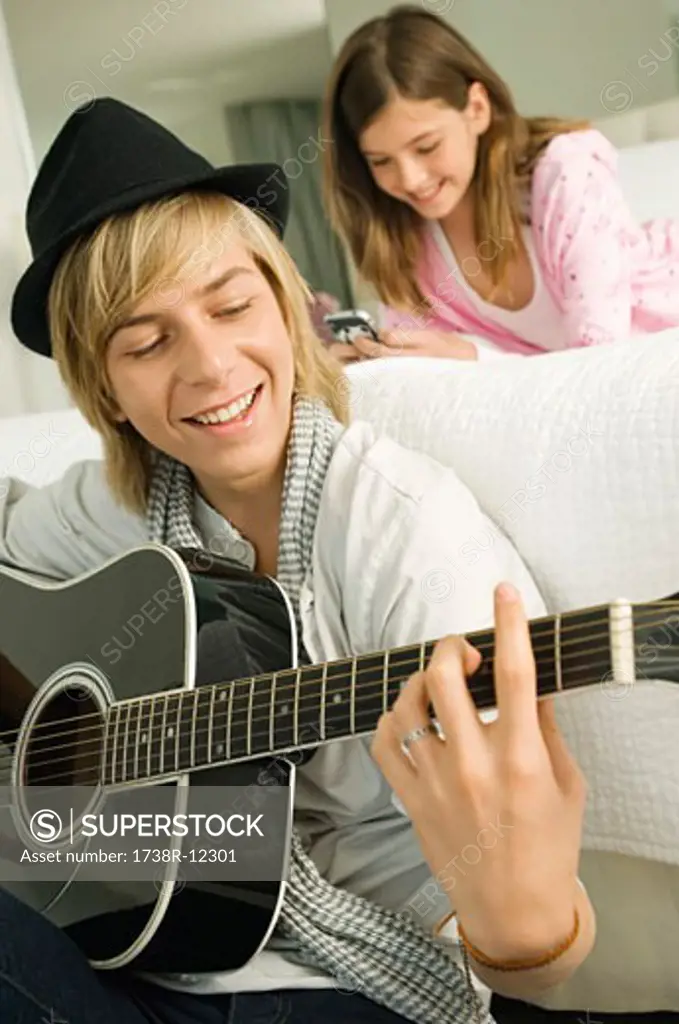 Teenage boy playing a guitar and his sister using a mobile phone behind him