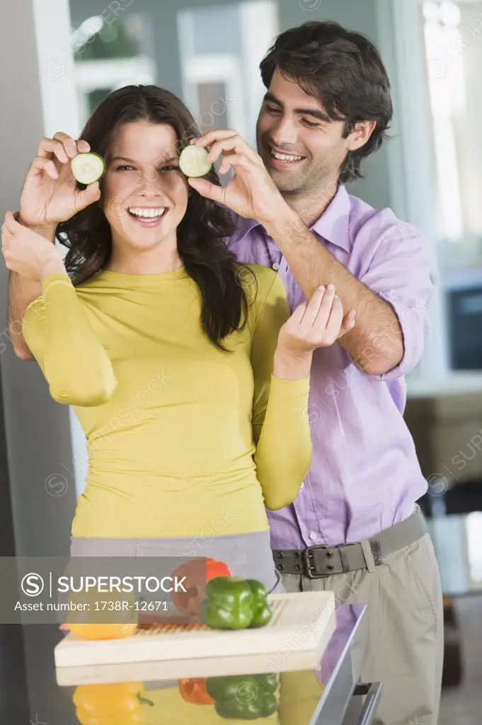 Man holding cucumber slices in front of a woman in the kitchen