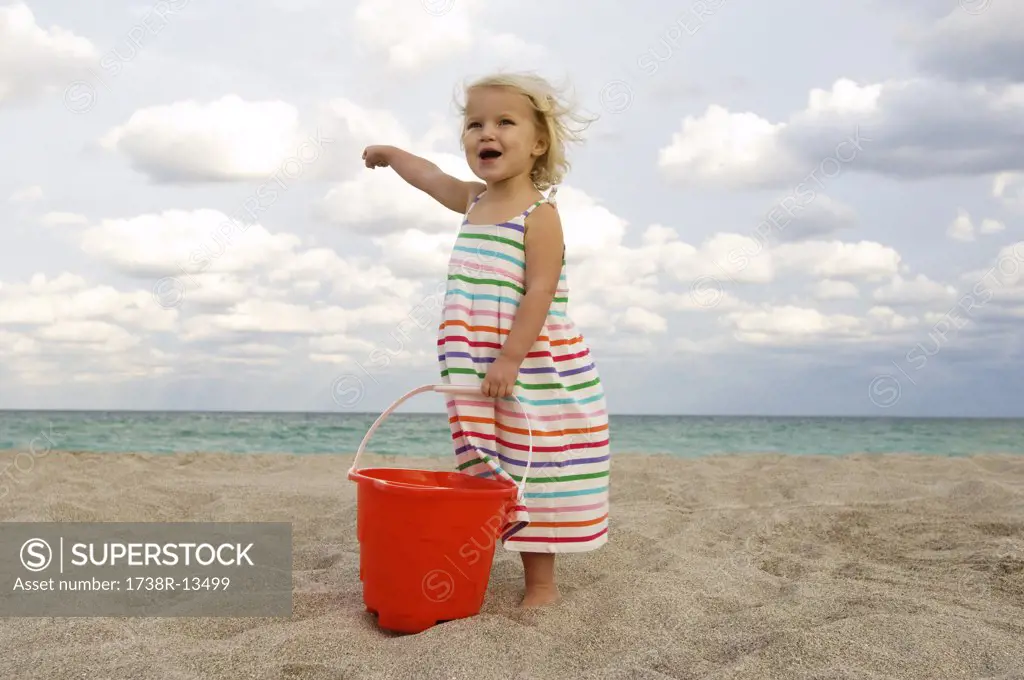 Girl holding a sand pail on the beach and pointing towards the sea