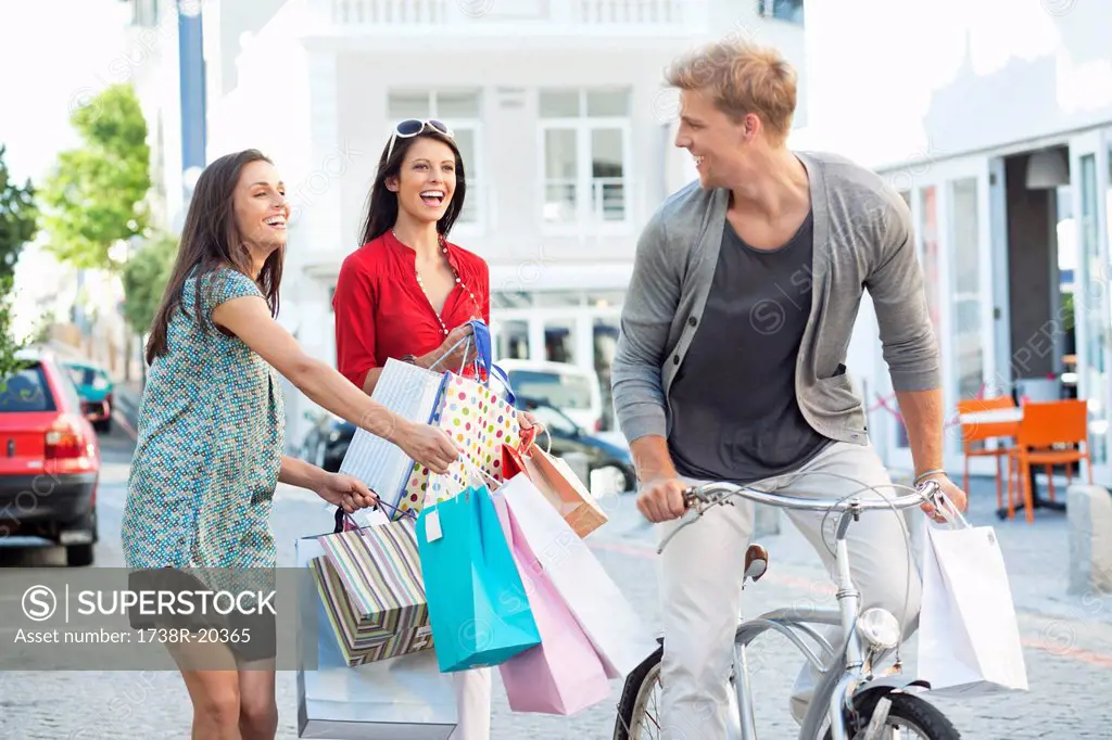 Young man cycling with two women running after him with shopping bags