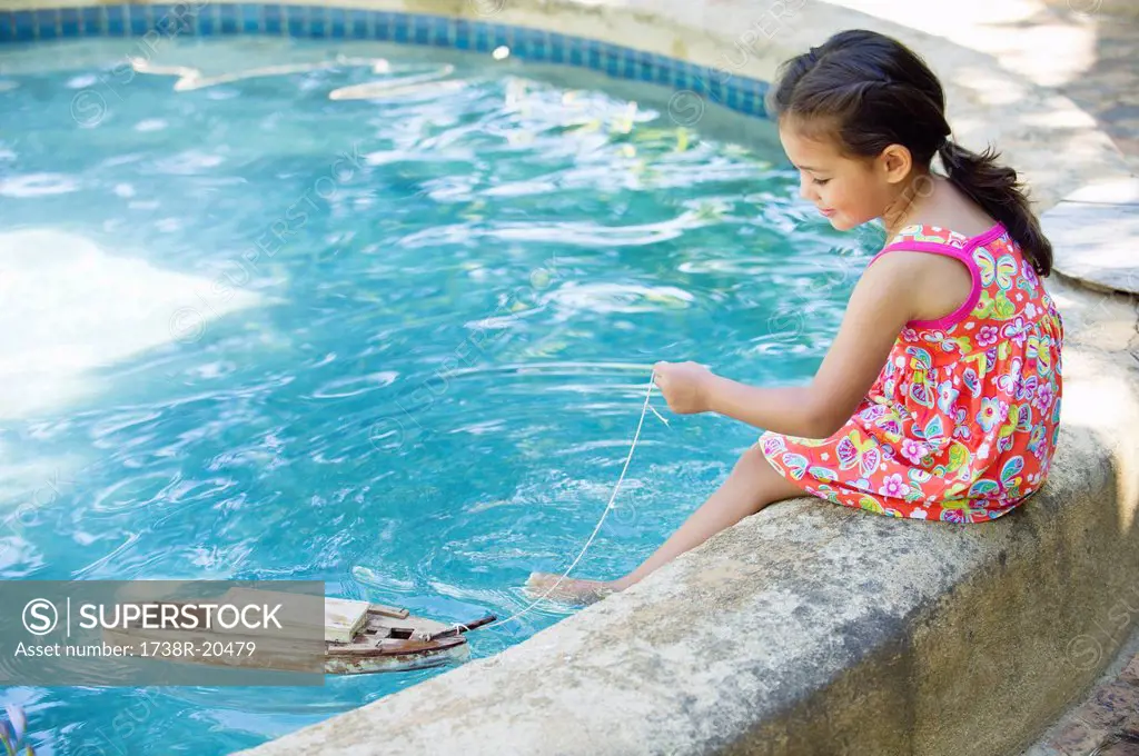 Girl sitting at edge of swimming pool looking at toy boat in water