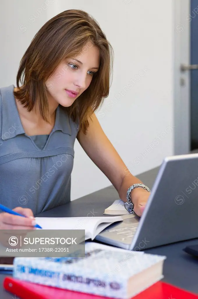 Beautiful woman writing in book while using laptop in classroom