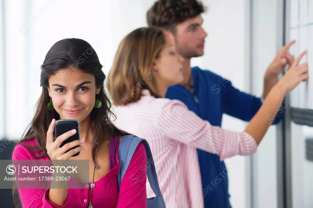Portrait of university students using a mobile phone while friends checking for test results in bulletin board