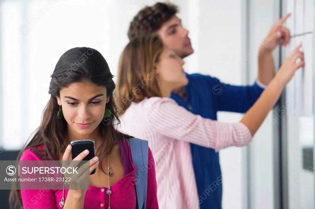 University students checking for test results in bulletin board while woman checking text message on mobile phone