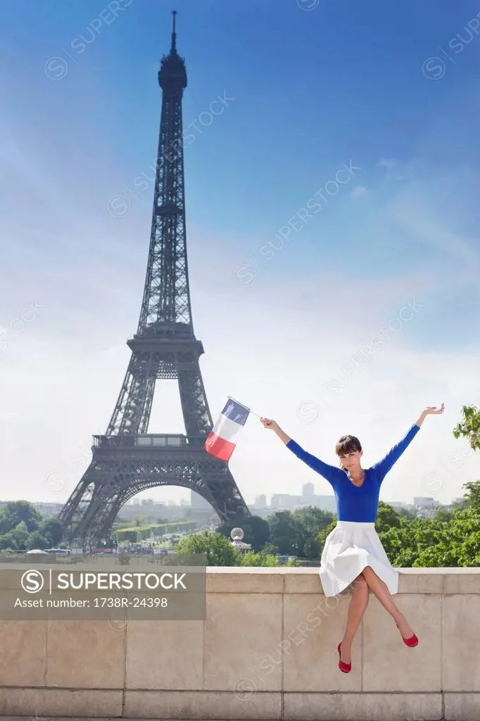 Woman holding a French flag sitting on a stone wall with the Eiffel Tower in the background, Paris, Ile_de_France, France