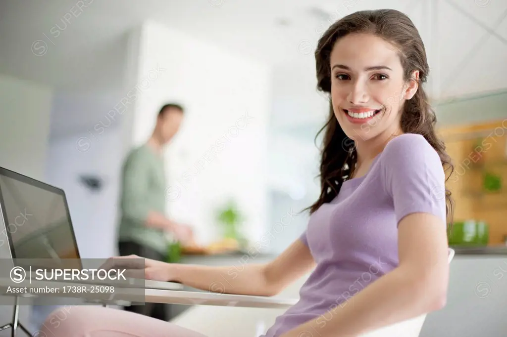 Woman using a laptop with her husband preparing food in the background