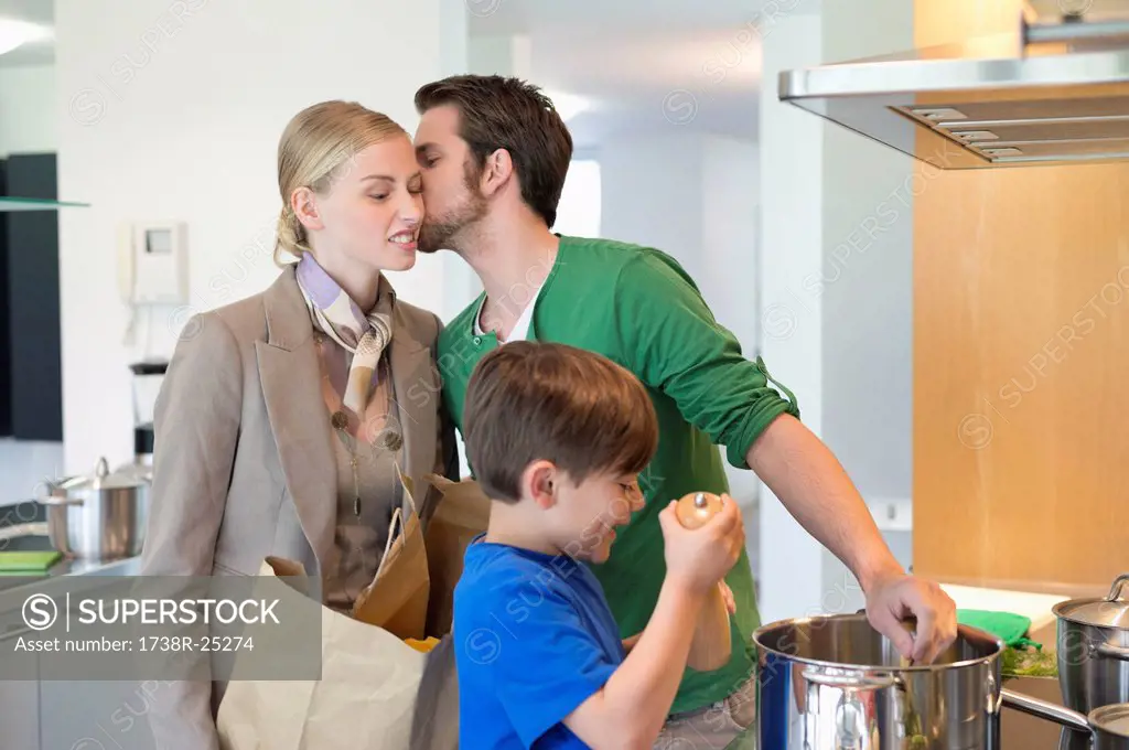 Man kissing his wife returning from shopping and son cooking in the kitchen
