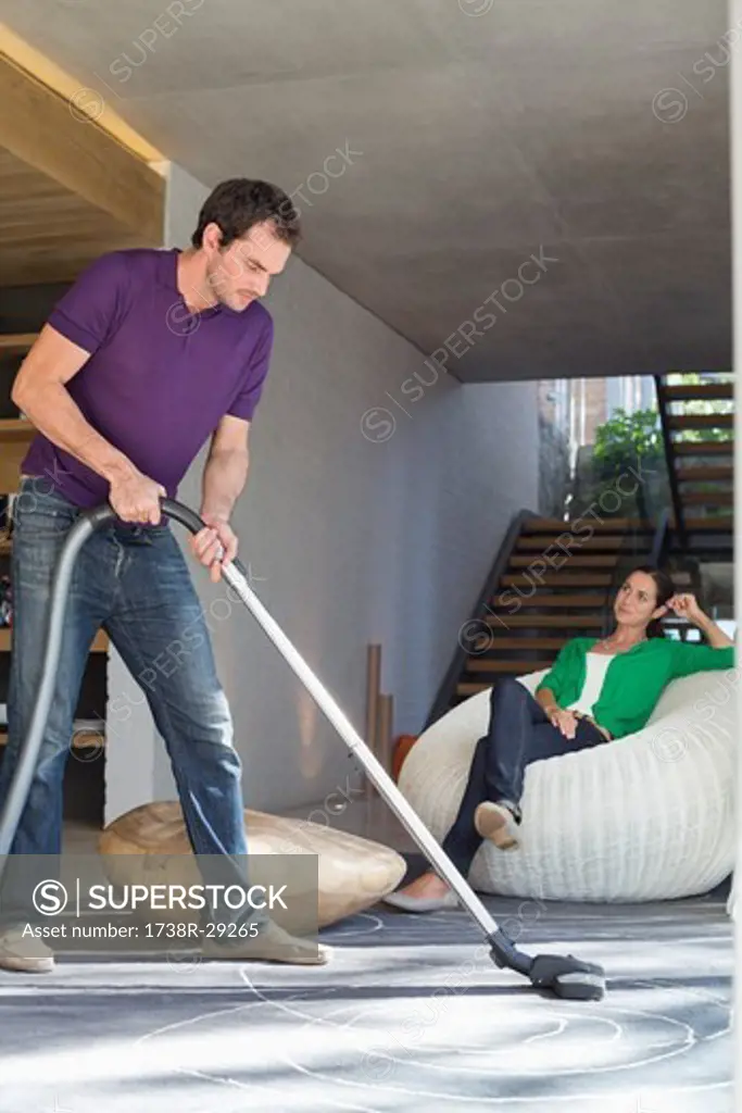 Man cleaning house with a vacuum cleaner with his wife sitting on a seat