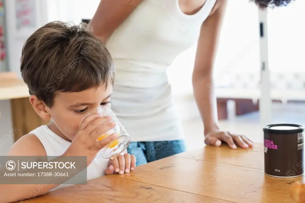 Boy drinking orange juice with his mother standing behind him
