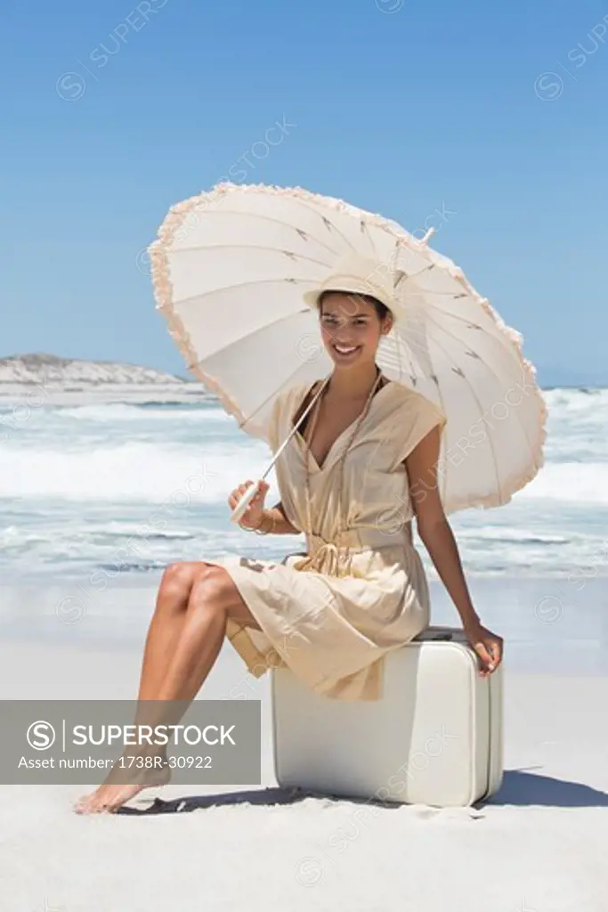 Beautiful woman sitting on a suitcase with an umbrella on the beach