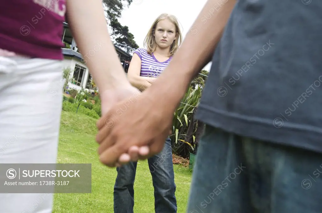 Teenagers holding hands, teen girl with arms crossed in background, outdoors