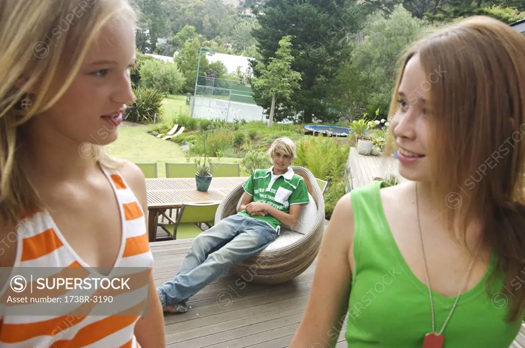 2 teenage girls looking at each other on a terrace, teenage boy sitting in background