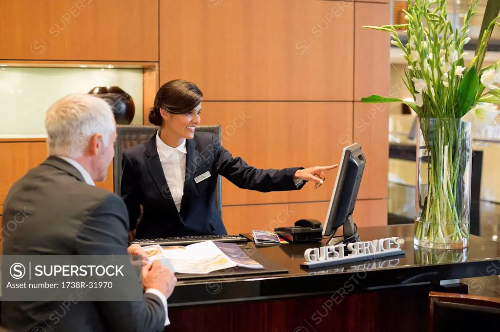 Receptionist pointing on a desktop pc with a businessman at the hotel reception counter