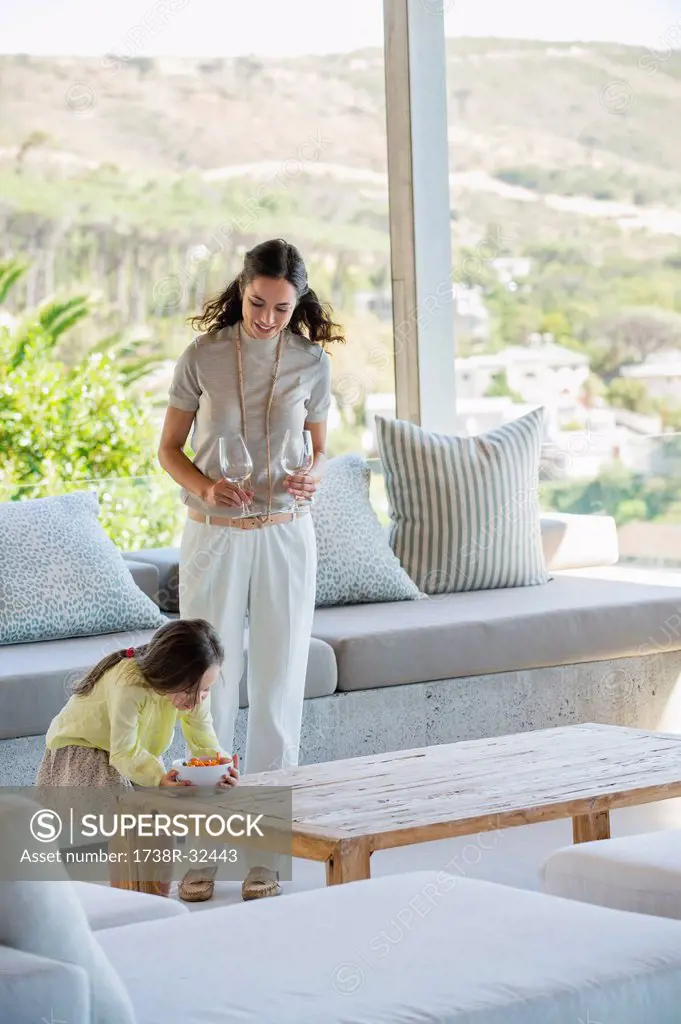 Girl putting a bowl of snacks on a table beside her mother holding glasses