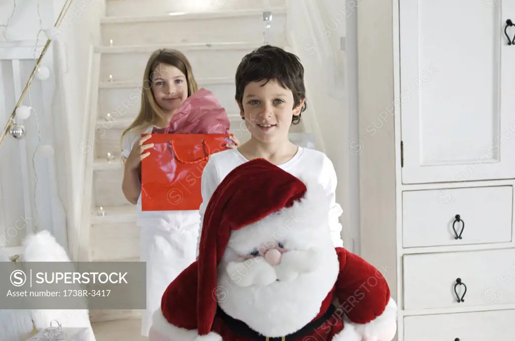 Christmas day, little boy holding a cuddly toy Santa Claus, looking at the camera, sister with present in background, indoors