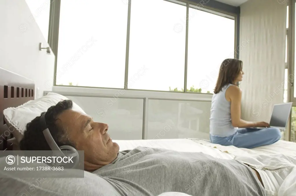 Couple in bedroom, man listening to music, woman using laptop, indoors