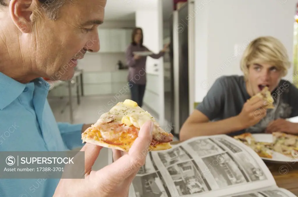 Father and teenager eating pizza, mother in background, indoors
