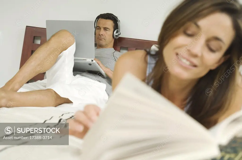 Couple on bed, woman reading, man using laptop, indoors