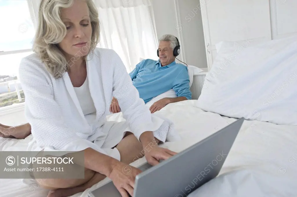 Woman using laptop in bedroom, man with headphones in background