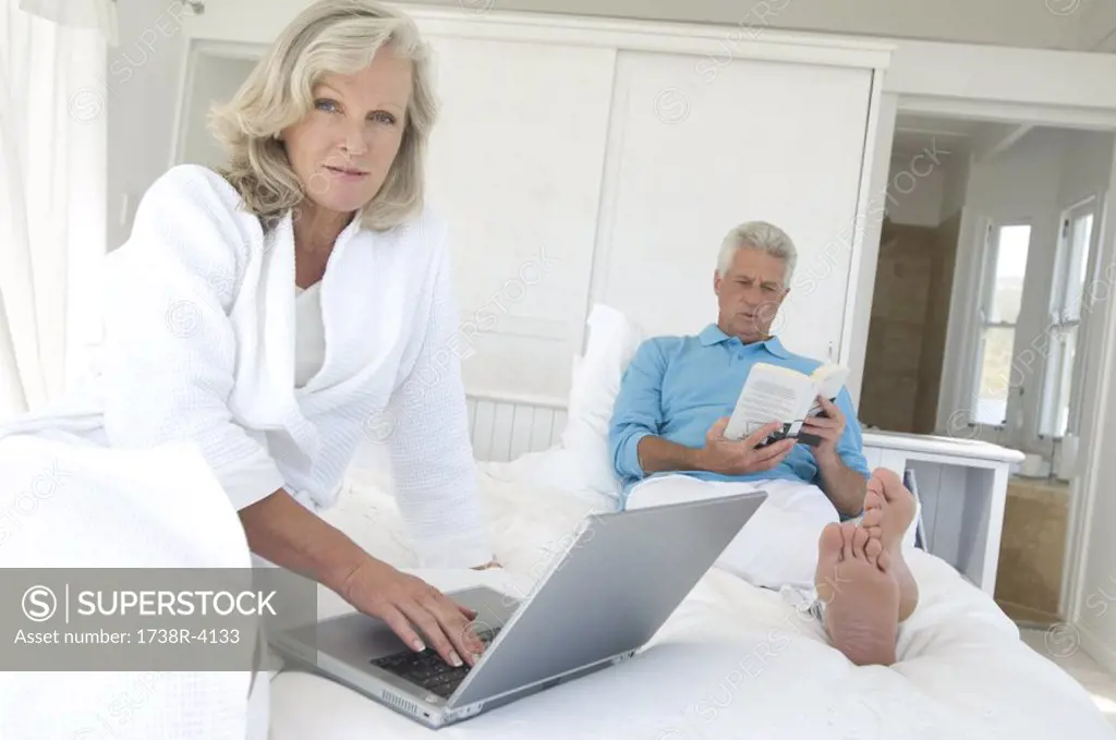 Couple in bed, man reading, woman using laptop