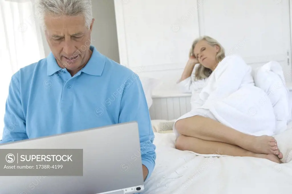 Couple in bedroom, man using laptop, woman lying in background