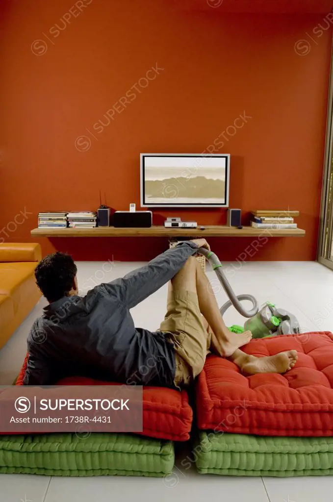 Man watching TV lying on cushions, vacuum cleaner in front of him
