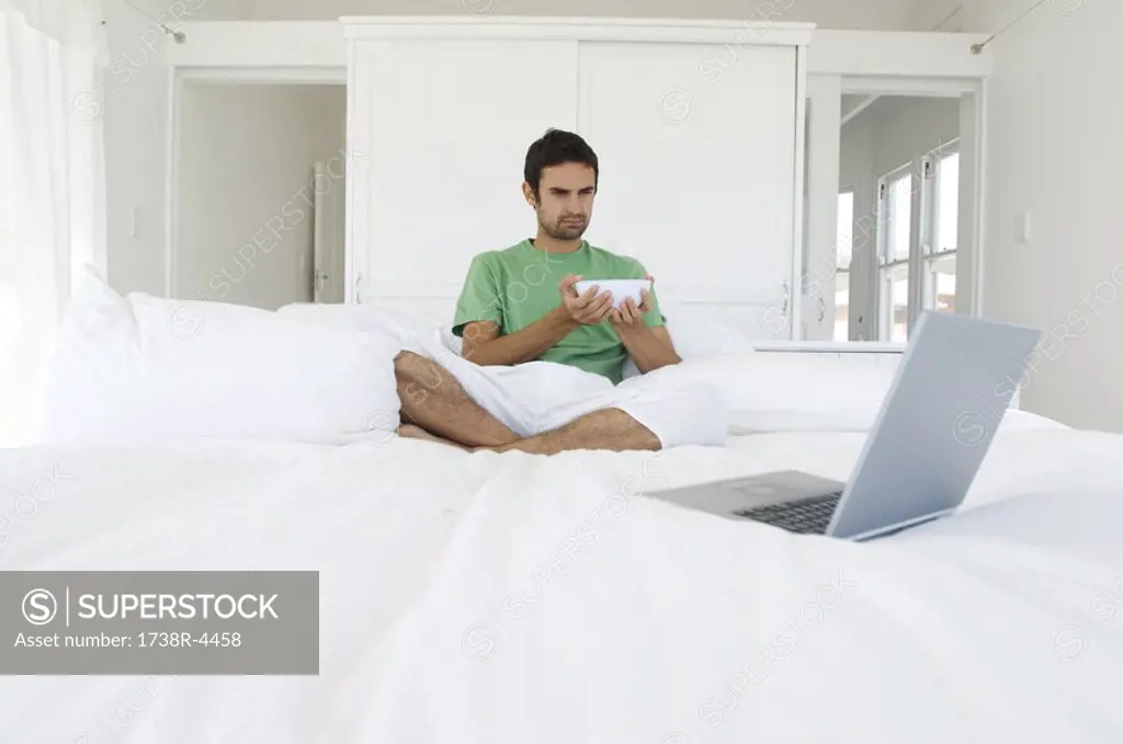 Young man sitting cross-legged on a bed, looking at laptop