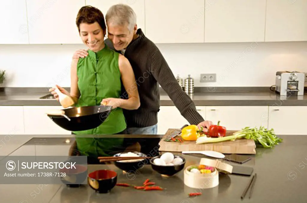 Mid adult woman preparing food with a mature man standing with arm around her in the kitchen