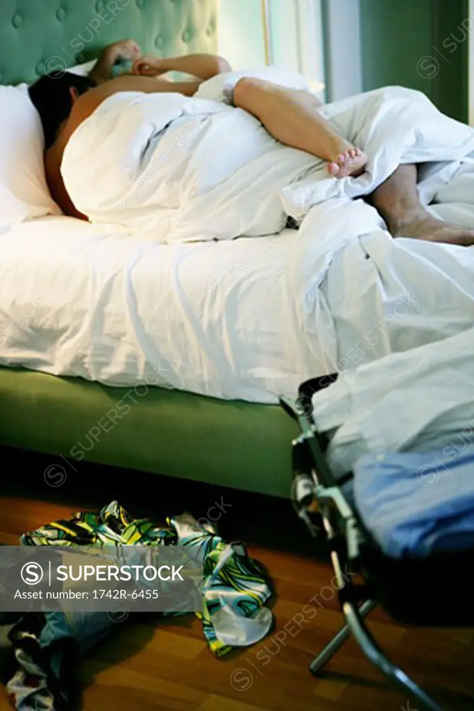 Couple under the covers in bed with clothes on floor. 