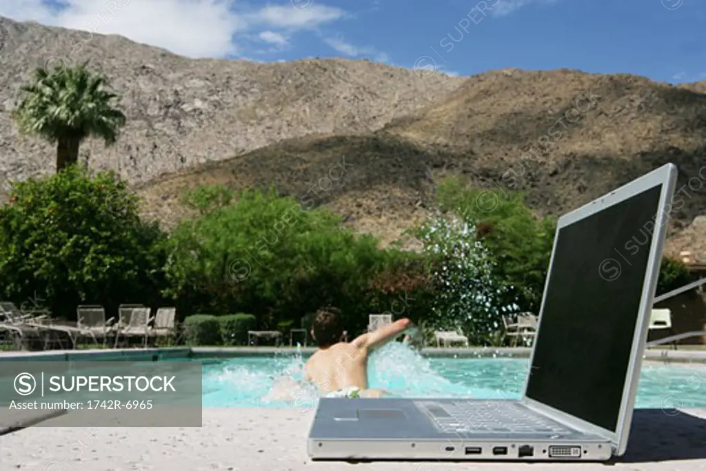 Laptop near pool with man swimming in background.