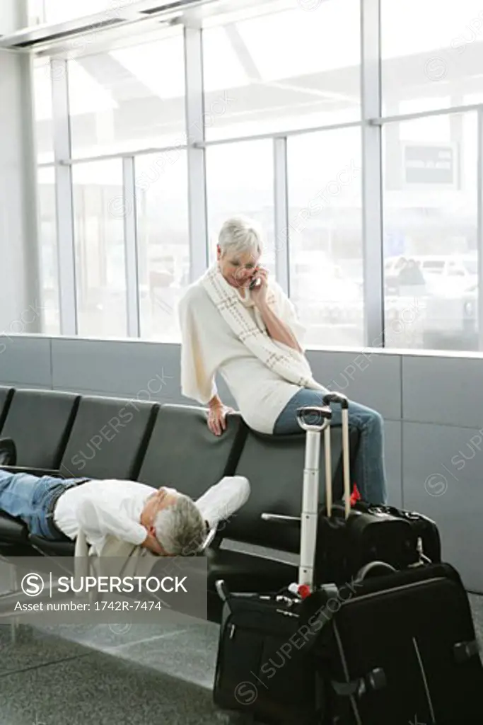 Mature woman on the phone at airport and mature man relaxing.