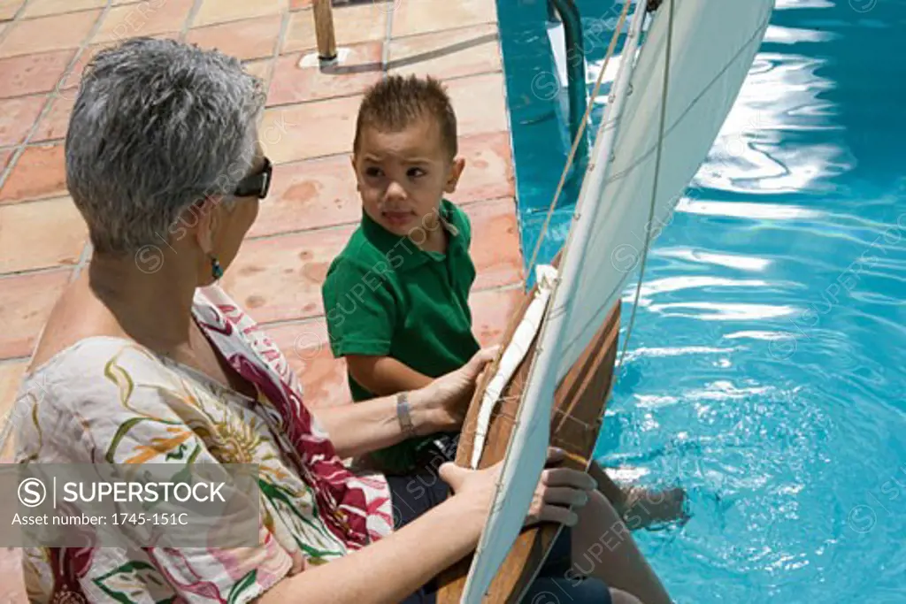 High angle view of a senior woman holding a sailboat with her grandson at poolside