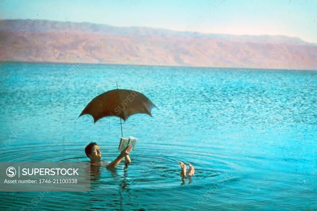 Colour photograph of a man floating in the Dead Sea whilst reading a book and holding an umbrella. Dated 1930