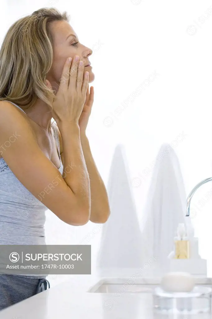 Woman standing at bathroom sink, touching face, looking at self