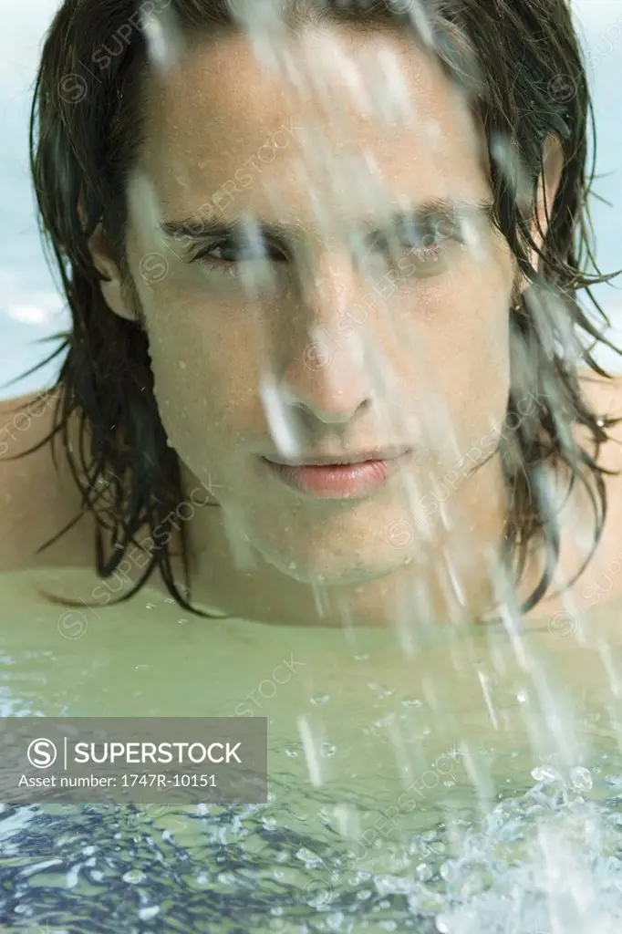 Man in swimming pool, looking at camera through splashing water