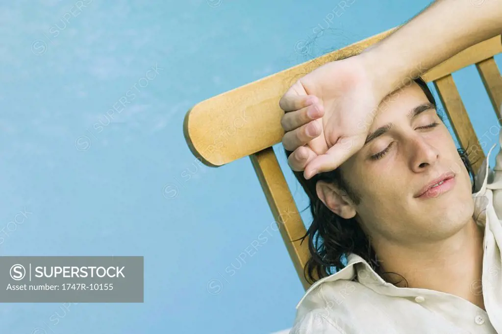 Man reclining in lounge chair beside pool, eyes closed, arm on head